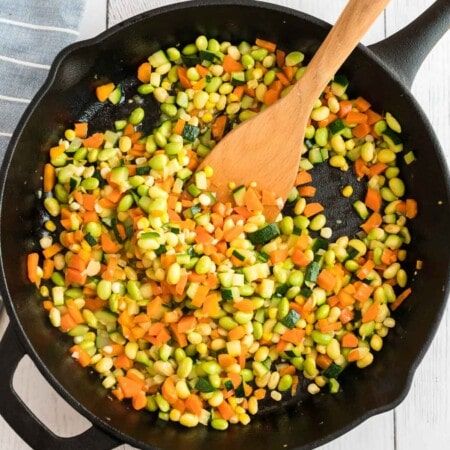 Una sartén de hierro fundido con una cuchara de madera y un montón de verduras picadas para succotash de maíz