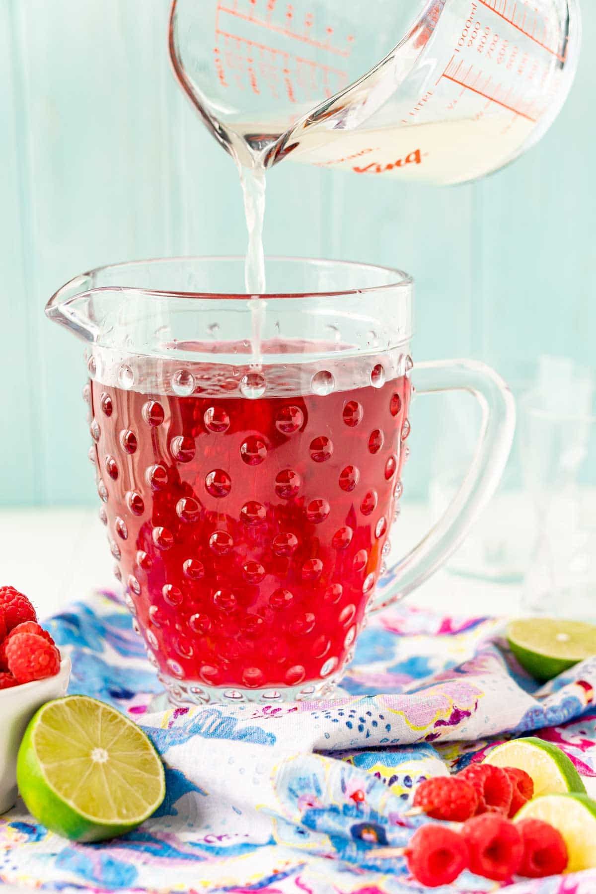 Una taza medidora vertiendo líquido transparente en una jarra de ponche de frambuesa roja