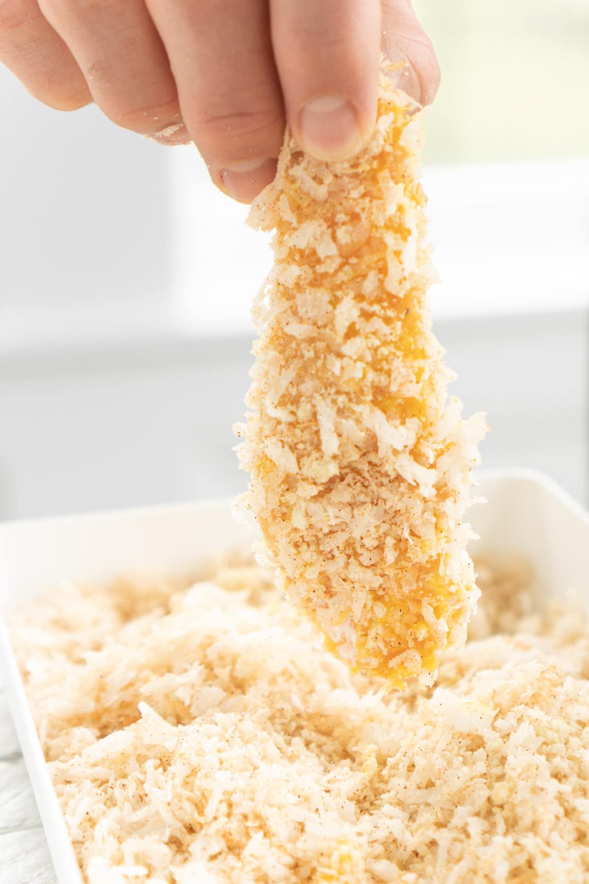 Χέρι που κρατά ψηλά ένα παναρισμένο πατατό κοτόπουλο καρύδας πάνω από το ψωμί κοτόπουλου καρύδας
