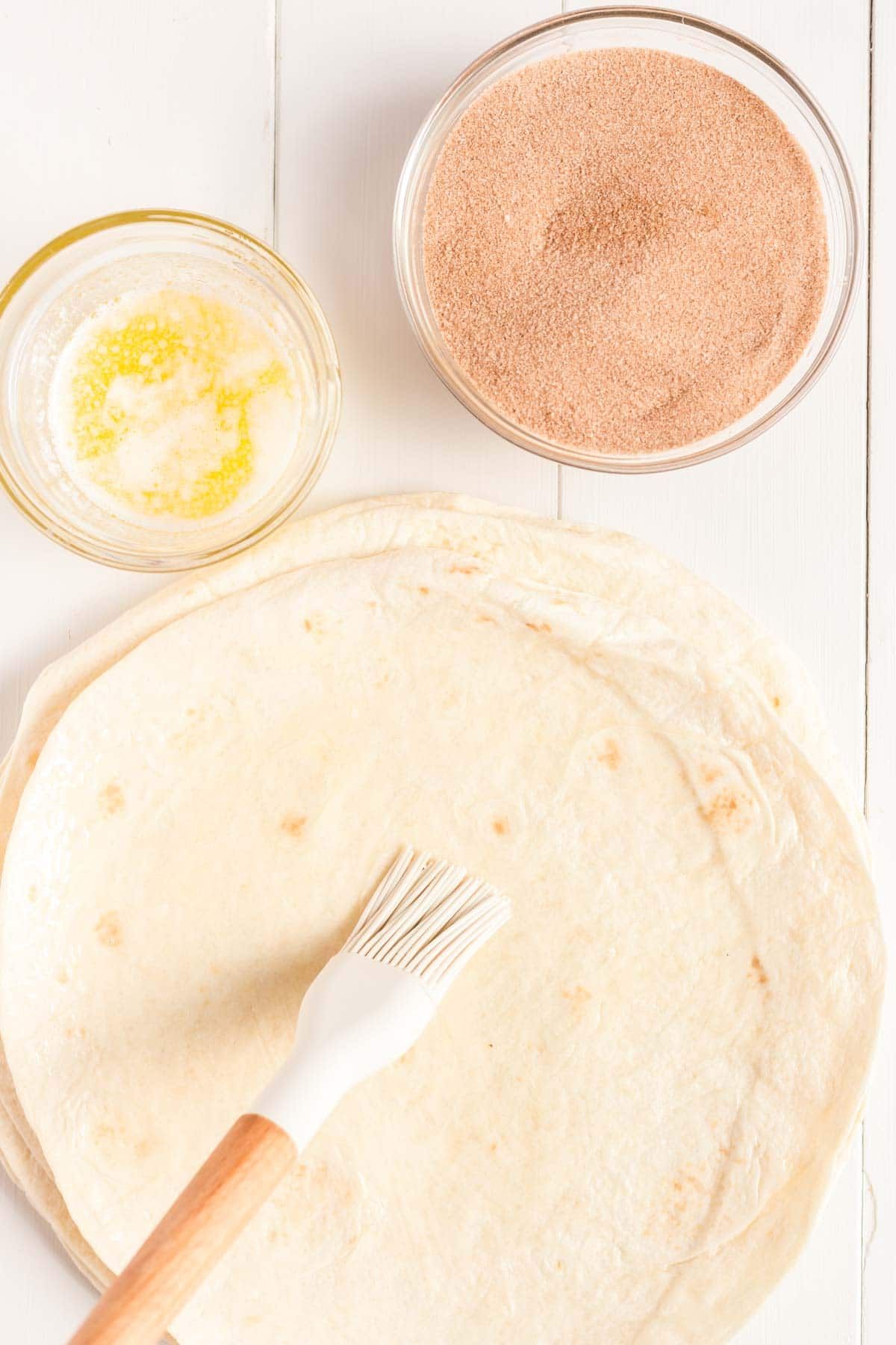 Silikonový kartáč s hromadou tortilly a miskou rozpuštěného másla a skořicového cukru