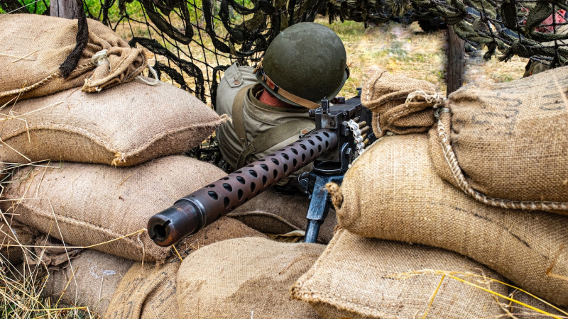   Persona con casco M1 mientras sostiene una ametralladora y se sienta sobre un montón de sacos